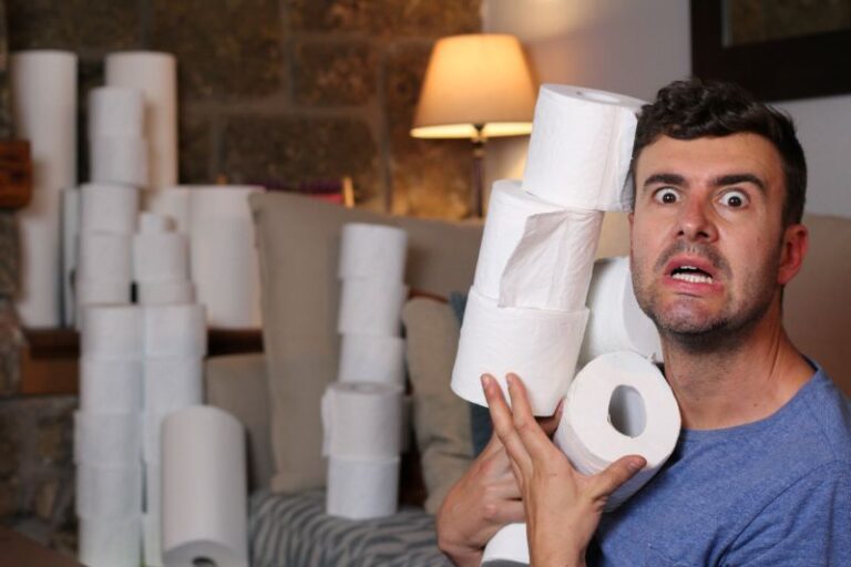 A surprised man sitting on a couch surrounded by stacks of toilet paper rolls, holding several rolls in his arms, visually representing confusion caused by unclear or exaggerated numbers.
